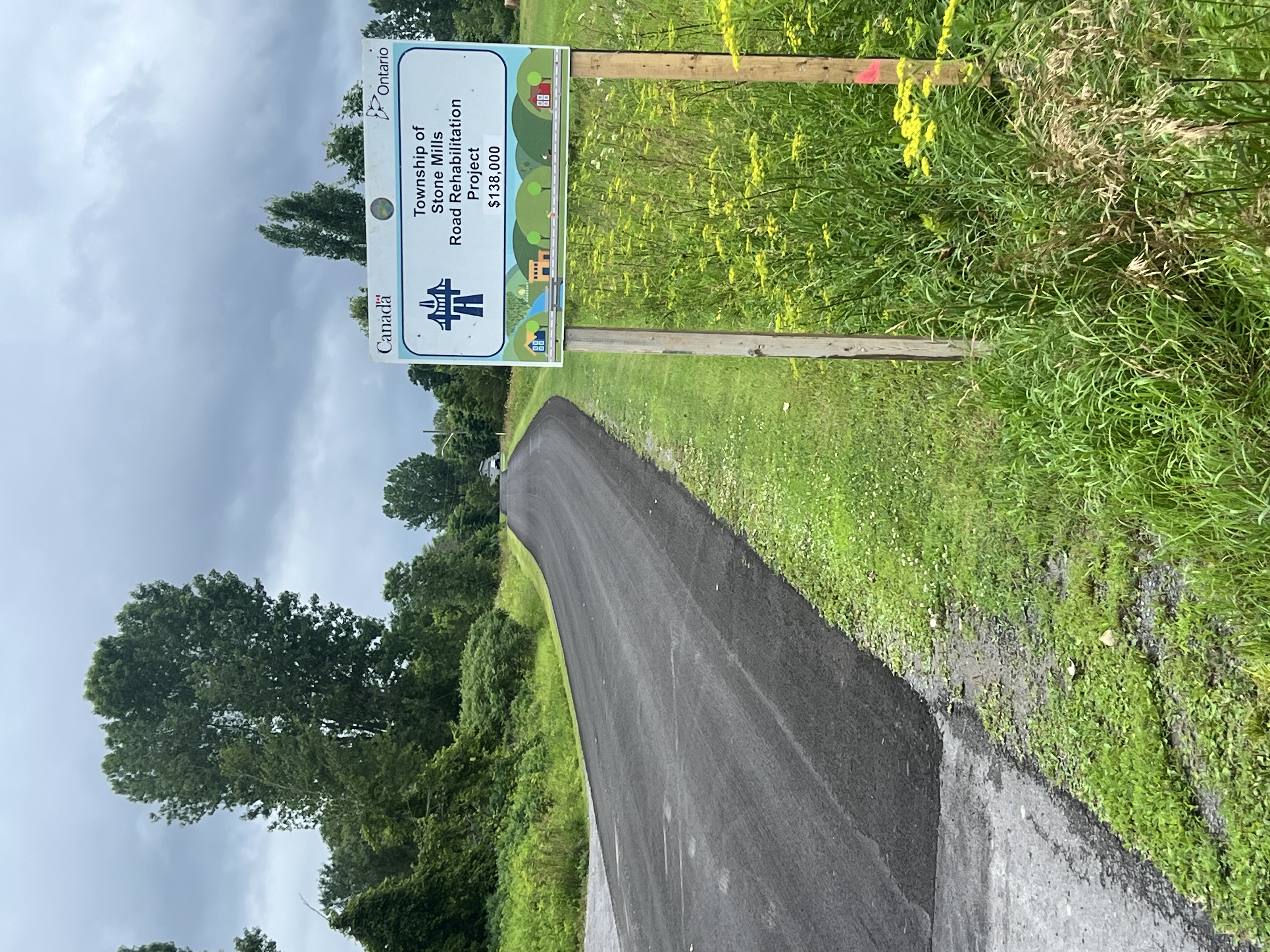 A rural roadway under an overcast sky with a project sign that says "Township of Stone Mills Road Rehabilitation Project".