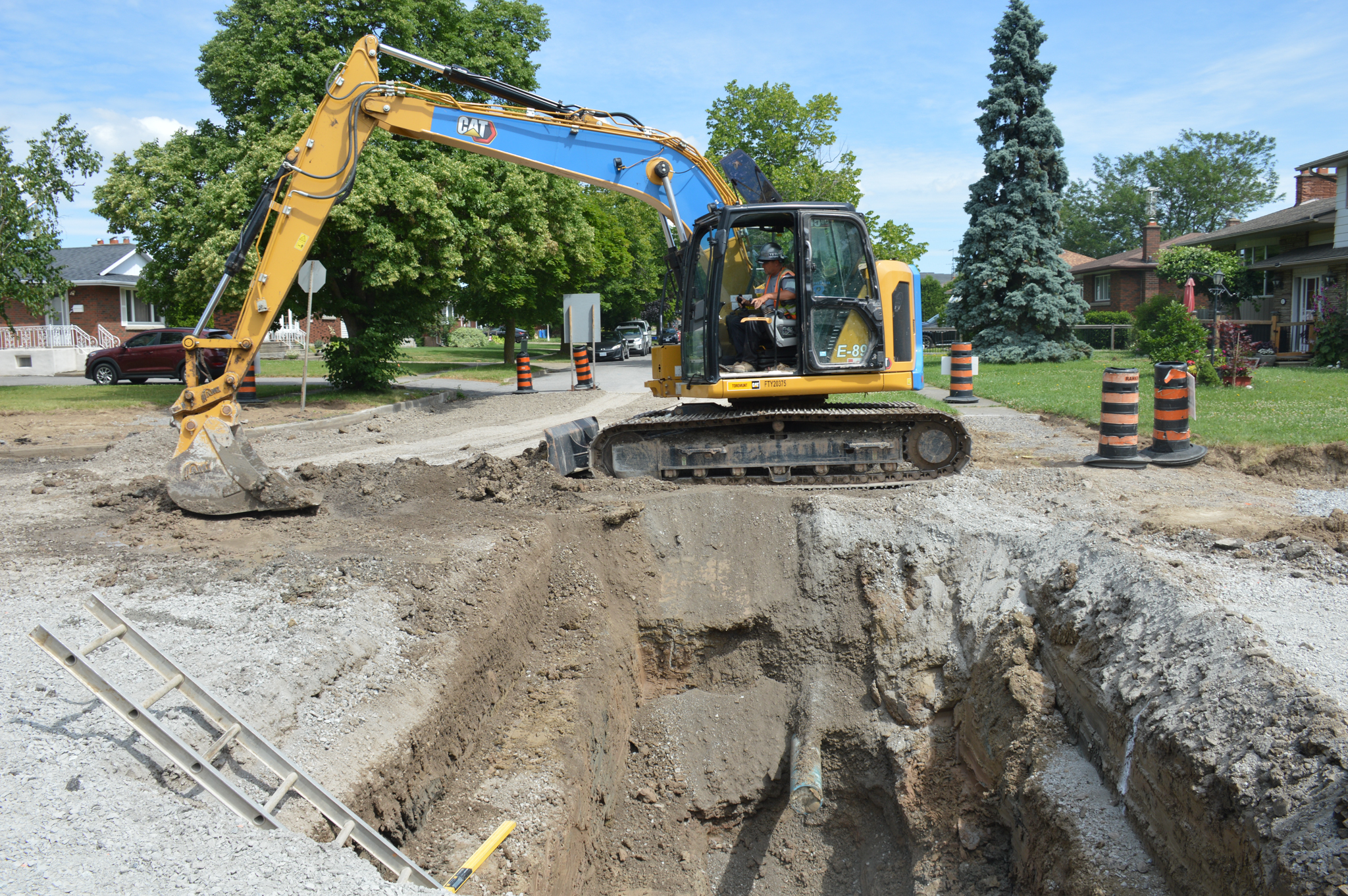 A construction worker in an excavator removes dirt from a large hole in the middle of an urban street.