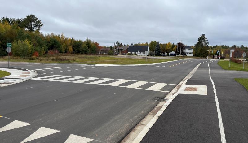 An asphalt roadway with a white painted crosswalk. There are houses in the background and an overcast sky. 